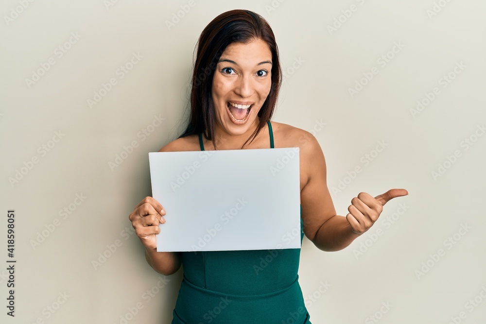Young latin woman holding blank empty banner pointing thumb up to the side smiling happy with open mouth Young latin woman holding blank empty banner pointing thumb up to the side smiling happy with open mouth