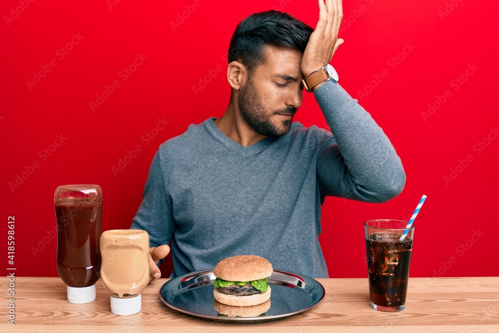 Handsome hispanic man eating a tasty classic burger and soda surprised with hand on head for mistake, remember error. forgot, bad memory concept. Handsome hispanic man eating a tasty classic burger and soda surprised with hand on head for mistake, remember error. forgot, bad memory concept.
