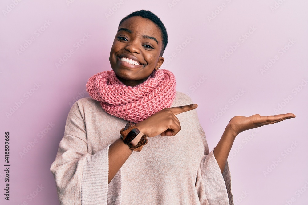 Young african american woman wearing winter scarf amazed and smiling to the camera while presenting with hand and pointing with finger. Young african american woman wearing winter scarf amazed and smiling to the camera while presenting with hand and pointing with finger.