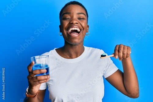 Young african american woman holding glass of mouthwash and toothbrush for fresh breath smiling and laughing hard out loud because funny crazy joke. photo
