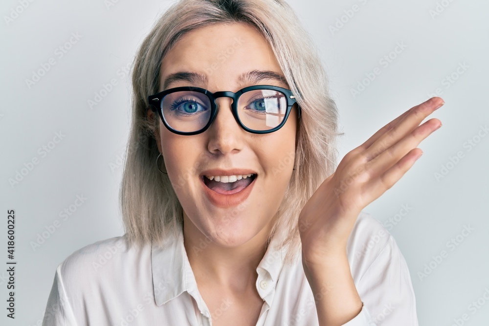 Young blonde girl wearing casual clothes and glasses celebrating achievement with happy smile and winner expression with raised hand Young blonde girl wearing casual clothes and glasses celebrating achievement with happy smile and winner expression with raised hand