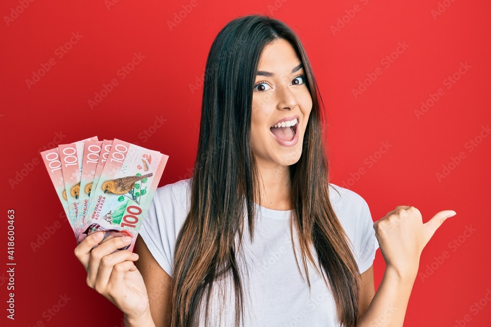 Young brunette woman holding 100 new zealand dollars banknotes pointing thumb up to the side smiling happy with open mouth Young brunette woman holding 100 new zealand dollars banknotes pointing thumb up to the side smiling happy with open mouth