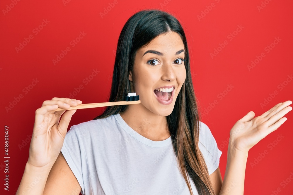 Young brunette woman holding toothbrush with toothpaste celebrating achievement with happy smile and winner expression with raised hand Young brunette woman holding toothbrush with toothpaste celebrating achievement with happy smile and winner expression with raised hand