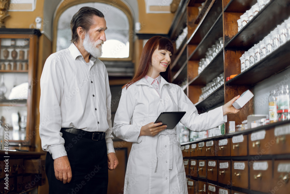 Young attractive Caucasian female pharmacist talking with pleasant senior male client in old vintage pharmacy, offering good safety medicine. Pharmaceutical care concept.. Young attractive Caucasian female pharmacist talking with pleasant senior male client in old vintage pharmacy, offering good safety medicine. Pharmaceutical care concept..