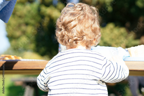 blond two-year-old boy with his back turned in a park sitting at a table.