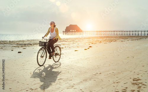 Middle-aged female dressed light summer clothes riding old vintage bicycle with front basket on the lonely low tide ocean white sand coast on Kiwengwa beach on Zanzibar island, Tanzania.