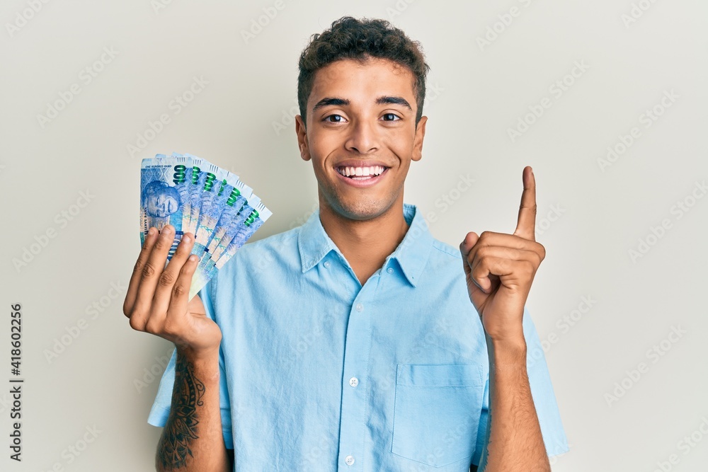 Young handsome african american man holding south african 100 rand banknotes smiling with an idea or question pointing finger with happy face, number one Young handsome african american man holding south african 100 rand banknotes smiling with an idea or question pointing finger with happy face, number one