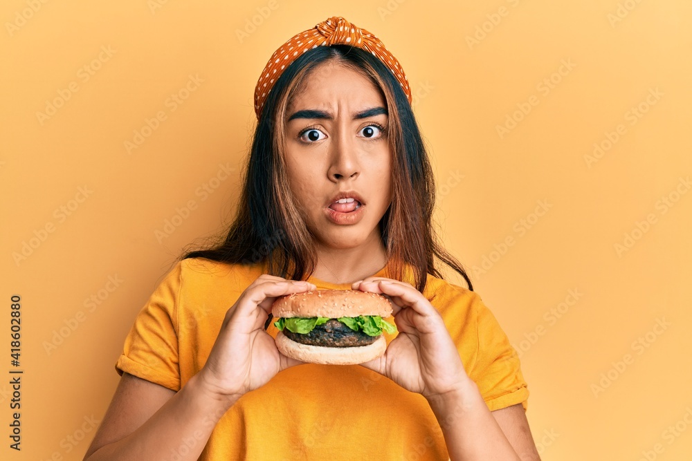 Young latin woman eating a tasty classic burger in shock face, looking skeptical and sarcastic, surprised with open mouth - obrazy, fototapety, plakaty 