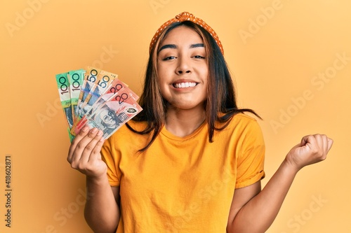 Young latin woman holding australian dollars banknotes screaming proud, celebrating victory and success very excited with raised arm