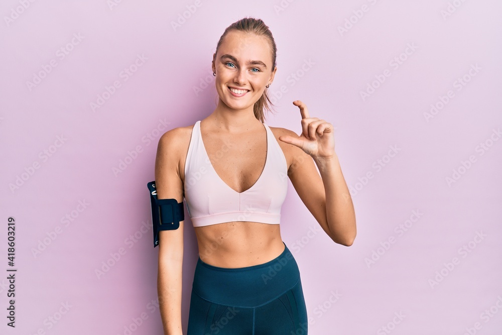 Beautiful blonde woman wearing sportswear and arm band smiling and confident gesturing with hand doing small size sign with fingers looking and the camera. measure concept.