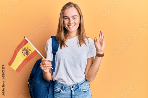 Beautiful blonde woman exchange student holding spanish flag doing ok sign with fingers, smiling friendly gesturing excellent symbol photo