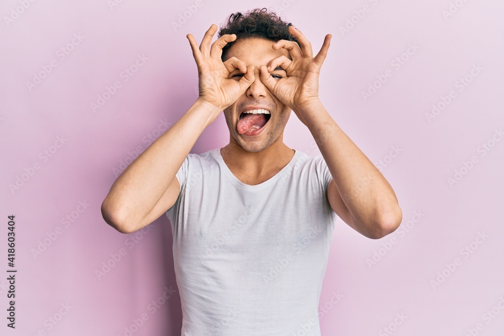 Young handsome man wearing casual white t shirt doing ok gesture like binoculars sticking tongue out, eyes looking through fingers. crazy expression. Young handsome man wearing casual white t shirt doing ok gesture like binoculars sticking tongue out, eyes looking through fingers. crazy expression.