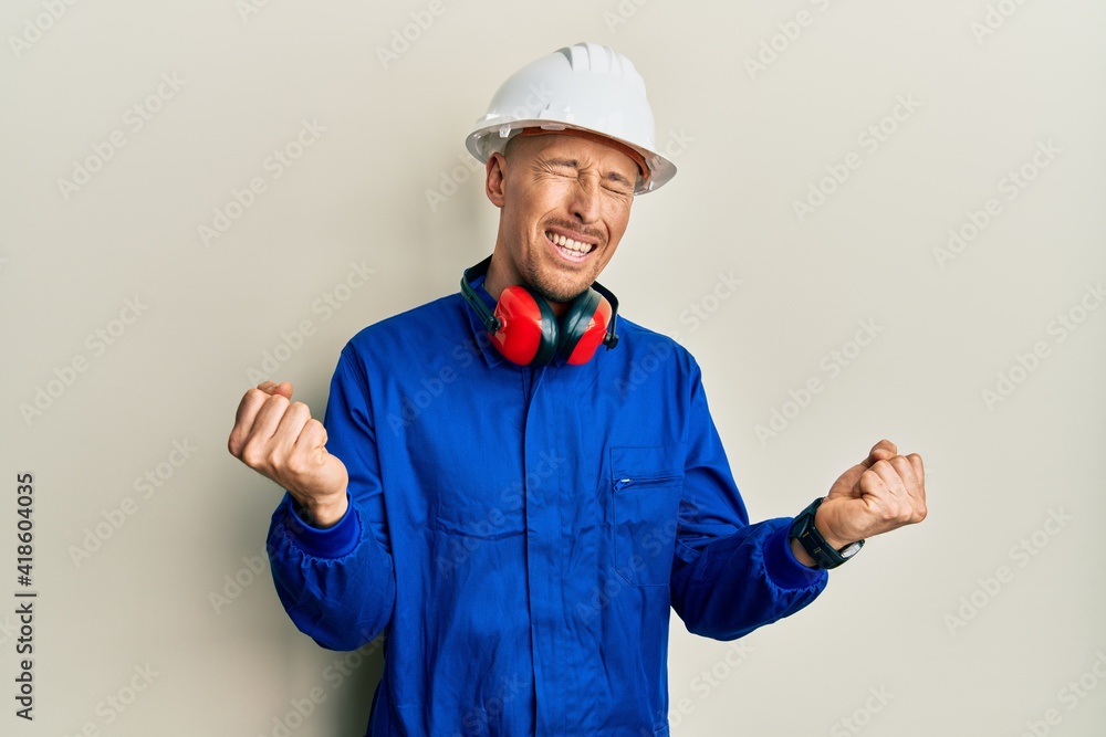 Bald man with beard wearing builder jumpsuit uniform and hardhat very happy and excited doing winner gesture with arms raised, smiling and screaming for success. celebration concept.