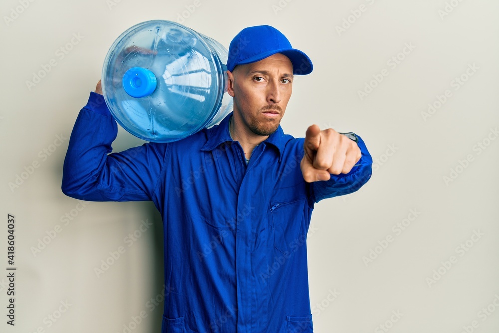 Bald courier man with beard holding a gallon bottle of water for delivery pointing with finger to the camera and to you, confident gesture looking serious
