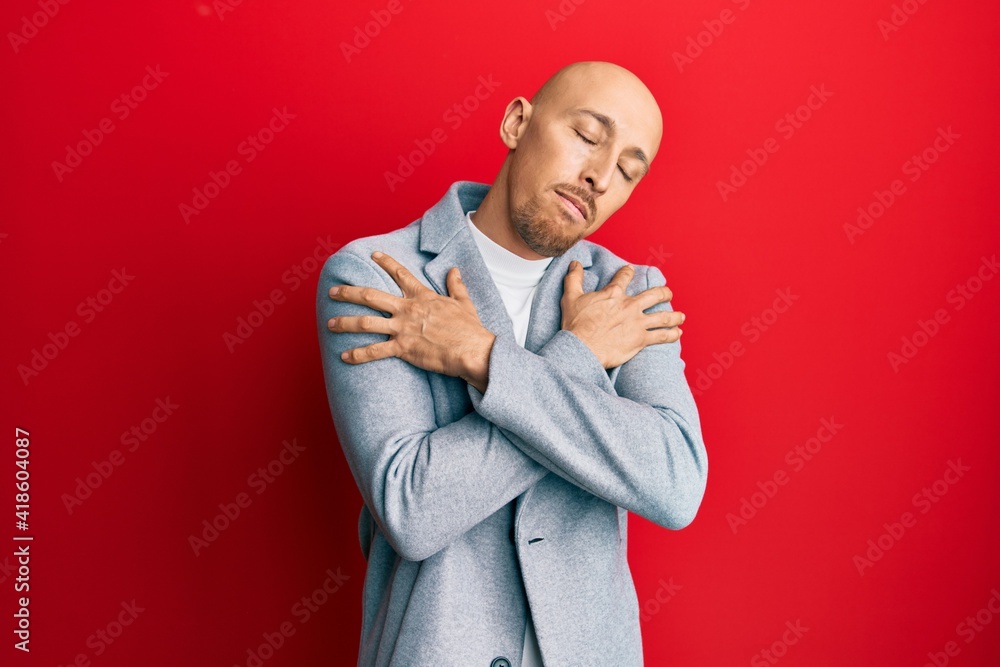 Bald man with beard wearing business jacket hugging oneself happy and positive, smiling confident. self love and self care