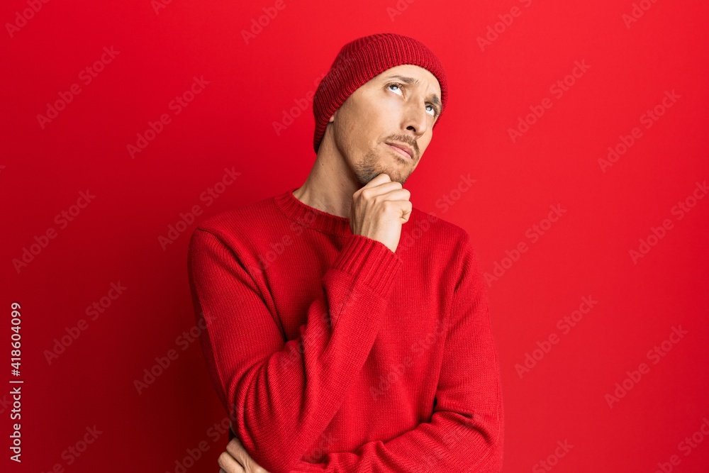 Bald man with beard wearing wool sweater and winter hat thinking concentrated about doubt with finger on chin and looking up wondering