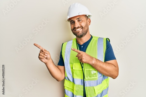 Handsome man with beard wearing safety helmet and reflective jacket smiling and looking at the camera pointing with two hands and fingers to the side. photo