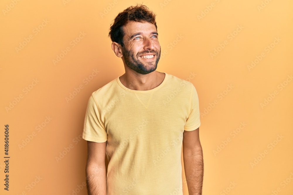 Handsome man with beard wearing casual yellow tshirt over yellow background looking away to side with smile on face, natural expression. laughing confident. Handsome man with beard wearing casual yellow tshirt over yellow background looking away to side with smile on face, natural expression. laughing confident.