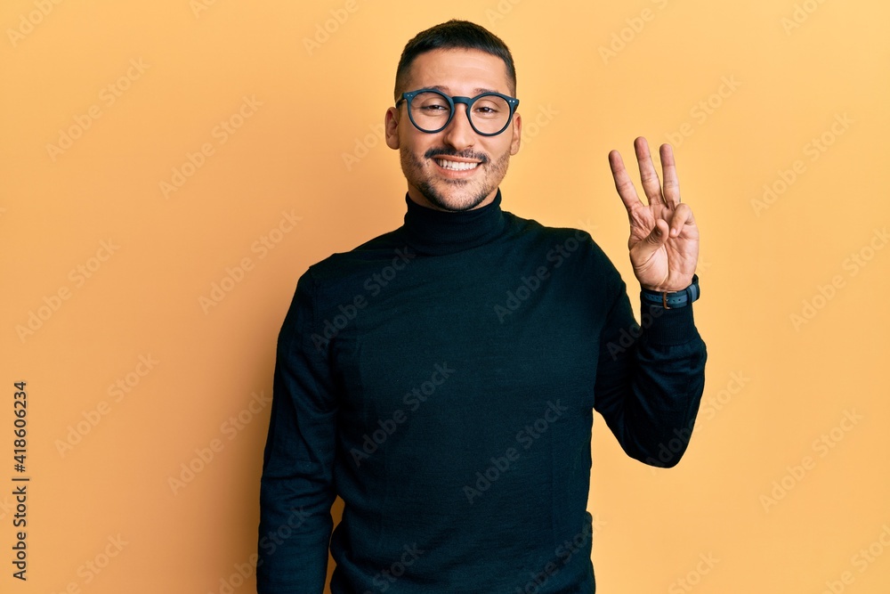 Handsome man with tattoos wearing turtleneck sweater and glasses showing and pointing up with fingers number three while smiling confident and happy. Handsome man with tattoos wearing turtleneck sweater and glasses showing and pointing up with fingers number three while smiling confident and happy.