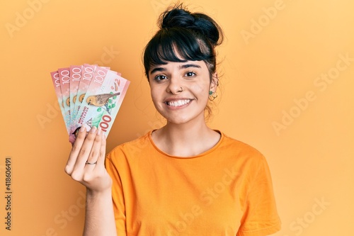 Young hispanic girl holding 100 new zealand dollars banknote looking positive and happy standing and smiling with a confident smile showing teeth