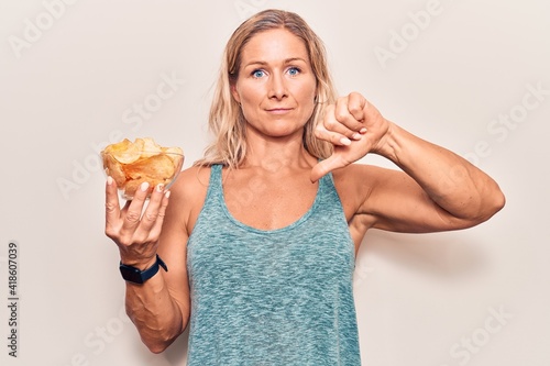 Middle age caucasian blonde woman holding potato chips with angry face, negative sign showing dislike with thumbs down, rejection concept