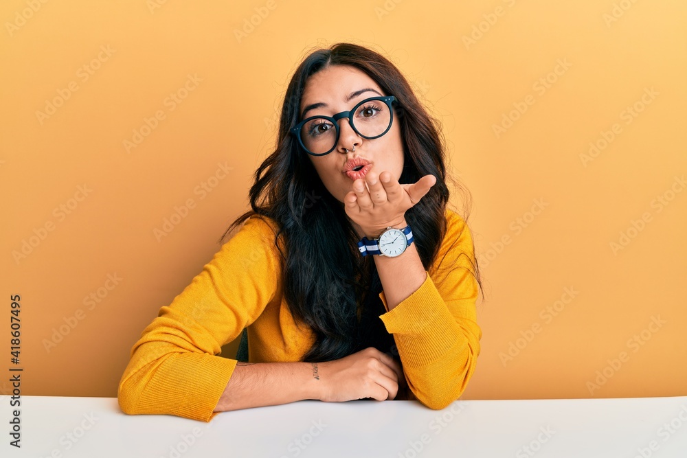 Beautiful brunette young woman wearing glasses and casual clothes sitting on the table looking at the camera blowing a kiss with hand on air being lovely and sexy. love expression. Beautiful brunette young woman wearing glasses and casual clothes sitting on the table looking at the camera blowing a kiss with hand on air being lovely and sexy. love expression.