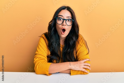 Beautiful brunette young woman wearing glasses and casual clothes sitting on the table afraid and shocked with surprise expression, fear and excited face.