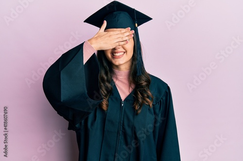 Young hispanic woman wearing graduation cap and ceremony robe smiling and laughing with hand on face covering eyes for surprise. blind concept.