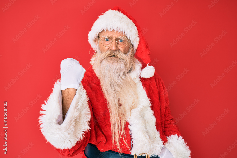 Old senior man with grey hair and long beard wearing traditional santa claus costume angry and mad raising fist frustrated and furious while shouting with anger. rage and aggressive concept. Old senior man with grey hair and long beard wearing traditional santa claus costume angry and mad raising fist frustrated and furious while shouting with anger. rage and aggressive concept.