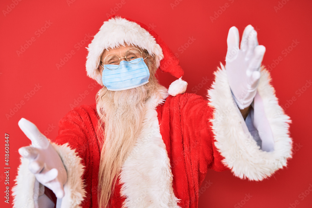 Old senior man wearing santa claus costume wearing safety mask looking at the camera smiling with open arms for hug. cheerful expression embracing happiness.