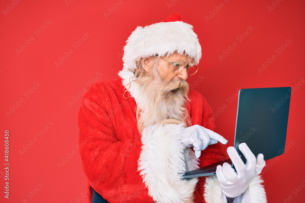 Naklejka premium Old senior man with grey hair and long beard wearing santa claus costume using laptop angry and mad screaming frustrated and furious, shouting with anger. rage and aggressive concept.