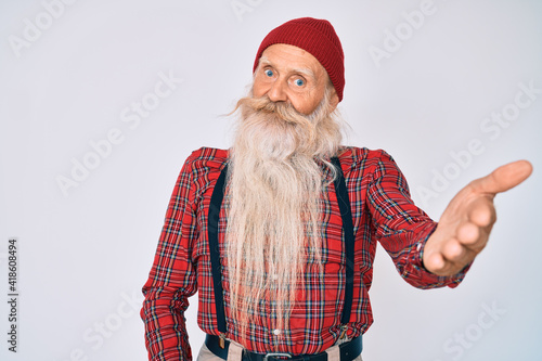 Old senior man with grey hair and long beard wearing hipster look with wool cap smiling friendly offering handshake as greeting and welcoming. successful business.
