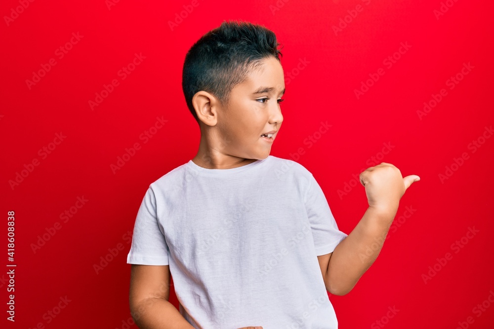 Little boy hispanic kid wearing casual white tshirt smiling with happy face looking and pointing to the side with thumb up. Little boy hispanic kid wearing casual white tshirt smiling with happy face looking and pointing to the side with thumb up.