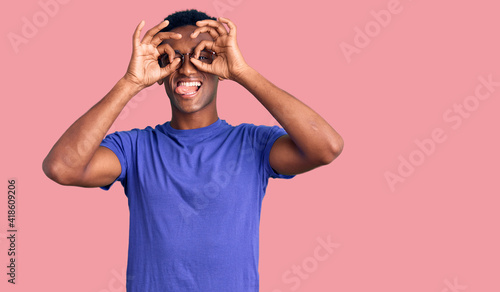 African handsome man wearing casual clothes and glasses doing ok gesture like binoculars sticking tongue out, eyes looking through fingers. crazy expression.