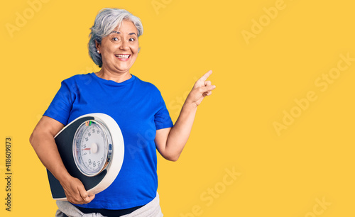 Senior woman with gray hair holding weight machine to balance weight loss smiling happy pointing with hand and finger to the side photo