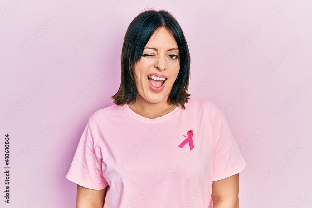 Fototapeta premium Young hispanic woman wearing pink cancer ribbon on t shirt winking looking at the camera with sexy expression, cheerful and happy face.