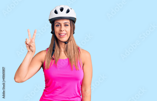 Young beautiful woman wearing bike helmet smiling with happy face winking at the camera doing victory sign. number two.