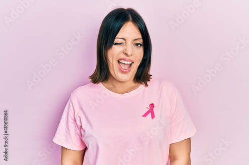 Young hispanic woman wearing pink cancer ribbon on t shirt winking looking at the camera with sexy expression, cheerful and happy face.