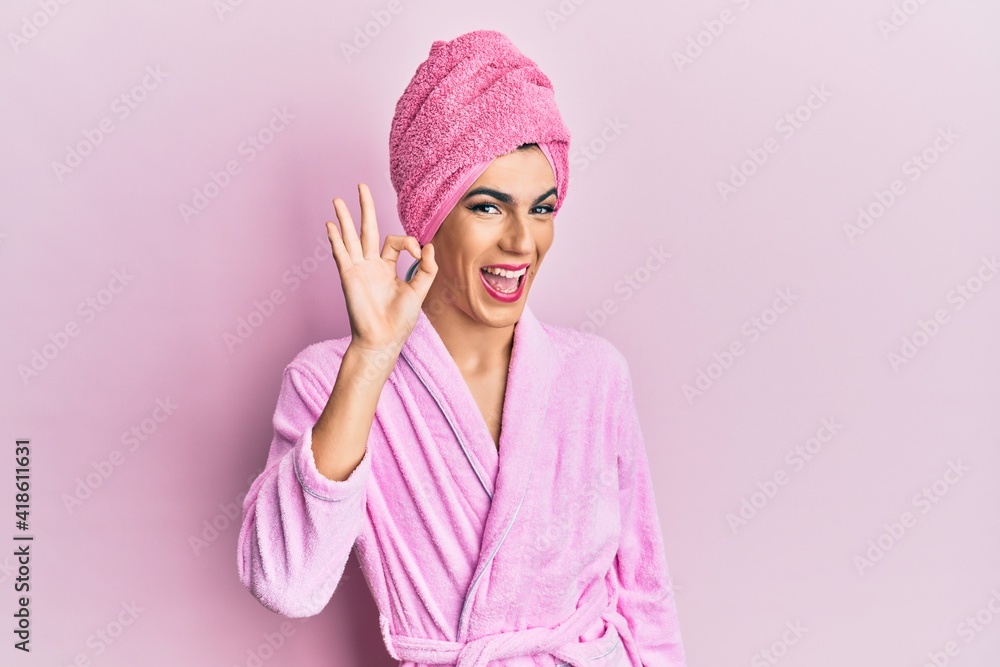 Young man wearing woman make up wearing shower towel on head and bathrobe smiling positive doing ok sign with hand and fingers. successful expression. Young man wearing woman make up wearing shower towel on head and bathrobe smiling positive doing ok sign with hand and fingers. successful expression.