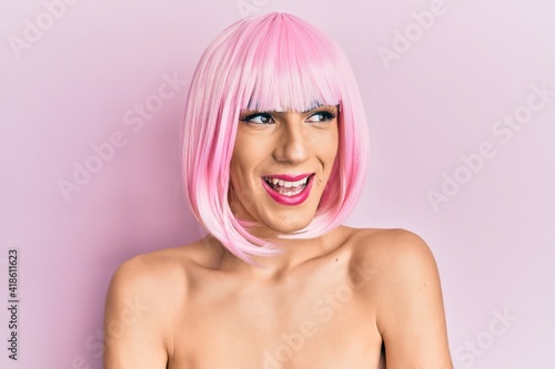 Young man wearing woman make up wearing pink wig smiling looking to the side and staring away thinking.
