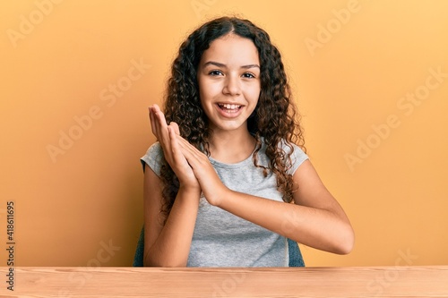 Teenager hispanic girl wearing casual clothes sitting on the table clapping and applauding happy and joyful, smiling proud hands together © Krakenimages.com