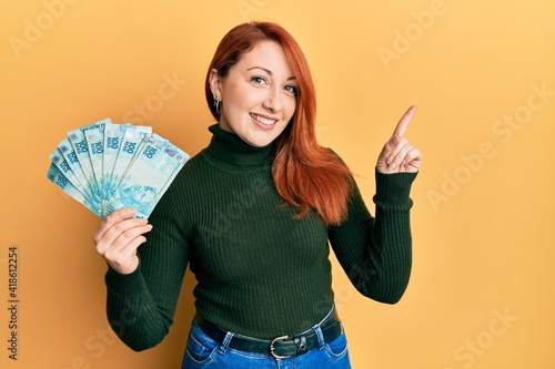Beautiful redhead woman holding 100 brazilian real banknotes smiling happy pointing with hand and finger to the side
