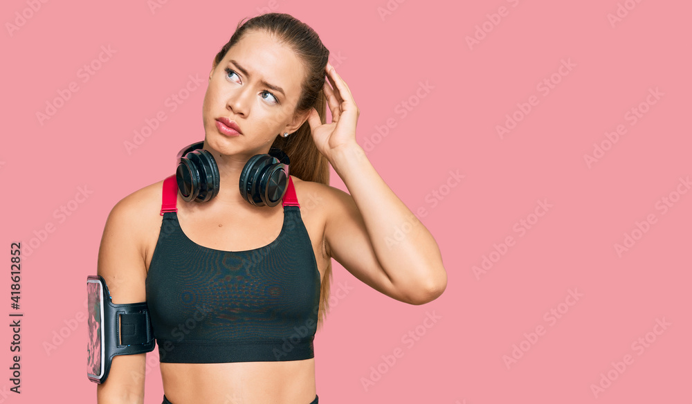 Beautiful blonde woman wearing gym clothes and using headphones confuse and wondering about question. uncertain with doubt, thinking with hand on head. pensive concept. Beautiful blonde woman wearing gym clothes and using headphones confuse and wondering about question. uncertain with doubt, thinking with hand on head. pensive concept.