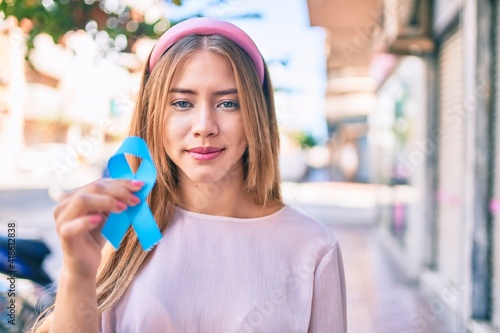 Young caucasian girl with serious expression holding blue cancer ribbon at the city.