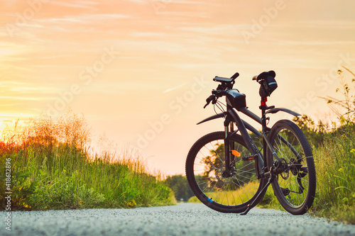 The mountain bike stands on a gravel bike path among green vegetation illuminated by the rays of the setting sun.