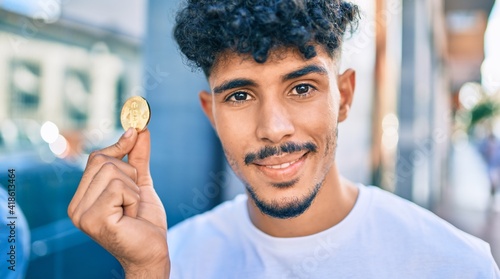 Young arab man smiling happy holding golden bitcoin walking at street of city.