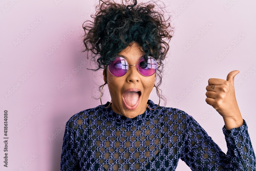 Young hispanic woman wearing cool sunglasses pointing thumb up to the side smiling happy with open mouth Young hispanic woman wearing cool sunglasses pointing thumb up to the side smiling happy with open mouth