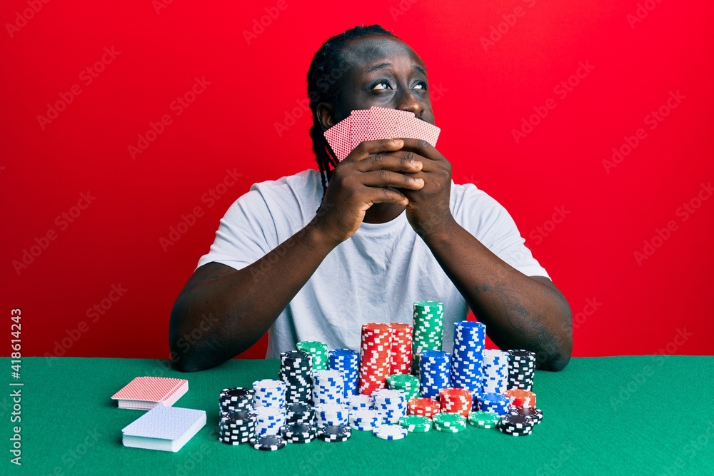 Handsome young black man playing gambling poker covering face with cards smiling looking to the side and staring away thinking. Handsome young black man playing gambling poker covering face with cards smiling looking to the side and staring away thinking.