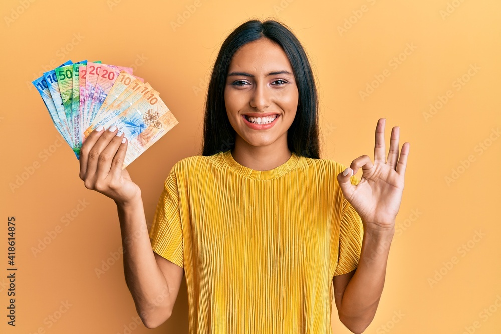 Young brunette woman holding swiss franc banknotes doing ok sign with fingers, smiling friendly gesturing excellent symbol Young brunette woman holding swiss franc banknotes doing ok sign with fingers, smiling friendly gesturing excellent symbol