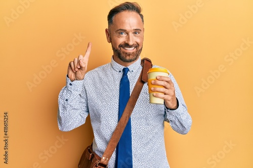 Handsome middle age business man wearing leather bag and drinking a take away cup of coffee smiling with an idea or question pointing finger with happy face, number one